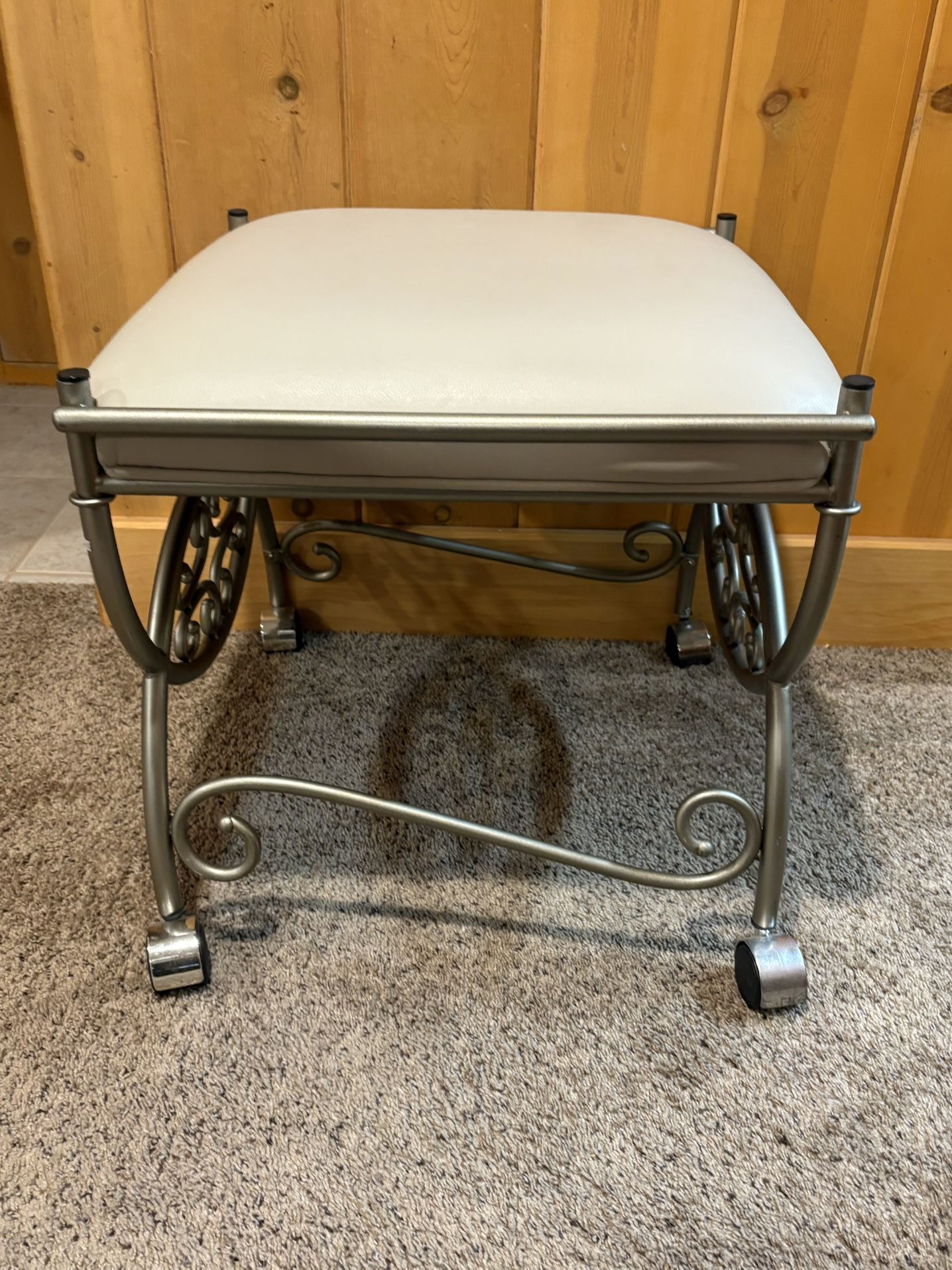 Victorian-style metal vanity stool with a silver finish, off-white upholstered seat, and casters for mobility.