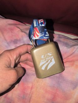 metal cowbell featuring the logo for "US Snowboarding".It includes a blue and red lanyard attached to the top for hanging