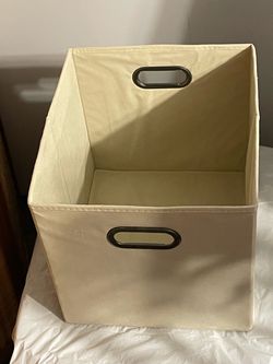 Cream fabric storage cube with dark oval carry handles on two sides. Sturdy open-top design with stitched edges. Good condition with light use.