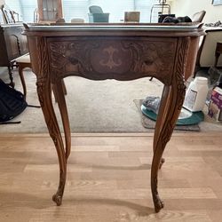 Ornate carved wooden gossip table with glass top and marquetry drawer.