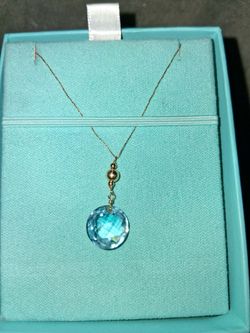 A round, checkerboard-cut blue gemstone pendant (such as blue topaz or aquamarine) styled as a delicate drop or briolette. It is accented with a polis