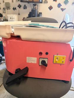 Pink pottery wheel machine has a metal turntable and a white plastic splash pan. This unit includes a foot pedal for speed control and a set of clay t