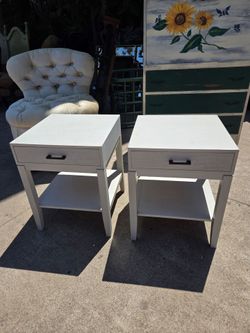 Two wooden nightstands with a brushed white finish, a front drawer, and an open shelf at the bottom.
