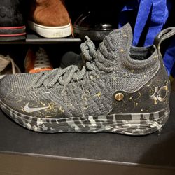 The shoes in the image are the Nike Zoom KD 11 "Gold Splatter" edition. 
