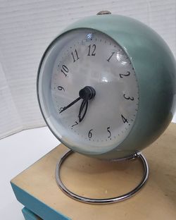 Mint-colored alarm clock is made of metal and glass. The clock face is protected by a glass cover with no cracks. It has a circular chrome-finished me