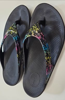 OOFOS Women's OOlala Limited recovery thong sandals in the Firefly colorway featuring a black base and a multi-color paint splatter pattern on the str