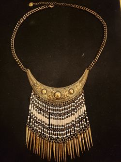 Ornate gold tone bib necklace with white tassel type goldtone pendant. Necklace is 17-in extending to 20-in.