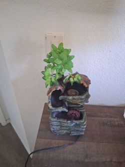 is an electric tabletop rockery water fountain featuring a tiered stone-look resin design, an integrated water wheel, and an artificial green tree.Des