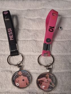 This image features two Inter Miami CF keychains inspired by Lionel Messi resting on a textured white cloth surface. The keychain on the left has a bl
