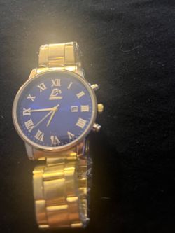 Elegant gold-tone stainless steel watch has a vibrant blue dial with Roman numeral hour markers. This timepiece features a date window at the 3 o'cloc