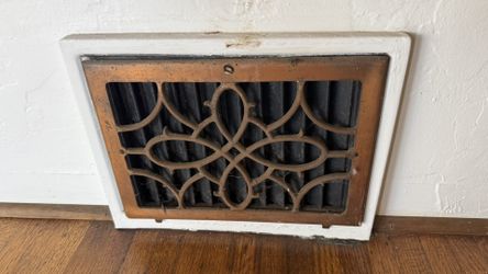 Ornate vent cover has a rustic bronze finish. It is made of cast iron with an intricate scrollwork design. Black louvered section is behind the decora