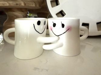 Hugging Smiley Face Mugs   Cute two white ceramic mugs. Mugs have smiley faces, one with pink blush. Mugs have arms to hug each other.