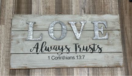 Rustic wooden decorative wall sign reading "LOVE Always Trusts" with the biblical reference 1 Corinthians 13:7. The design combines multi-textured ele