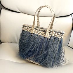 bolso de mano es un diseño artesanal, comúnmente elaborado con paja toquilla o rafia, y se caracteriza por sus llamativos flecos en tonos azules y un 