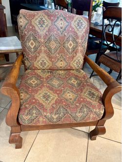 Sturdy wooden armchair has tapestry upholstery with red and gold geometric patterns. Frame is solid wood with curved armrests and decorative feet.