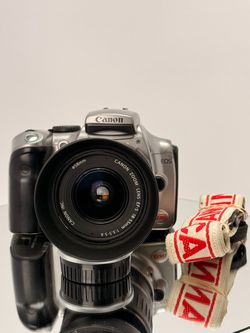 Canon EOS DSLR in silver and black body with Canon EF-S 18-55mm f/3.5-5.6 zoom lens attached. Camera strap included.