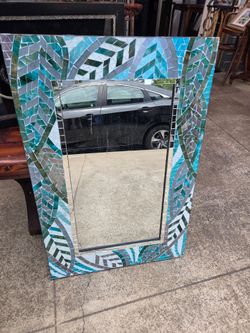 The wall mirror features a rectangular frame adorned with a handcrafted glass mosaic in shades of teal, blue, and gray.