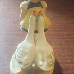 This charming figurine depicts a young girl with two geese, often shown with the geese stretching their necks as if looking for a treat. 