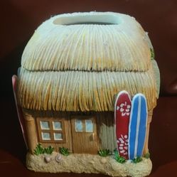 This is a Tiki Hut Tissue Box Cover Holder by Izsy. 
It is made of resin.
It features beach-themed elements like surfboards, plumeria flowers, and a p