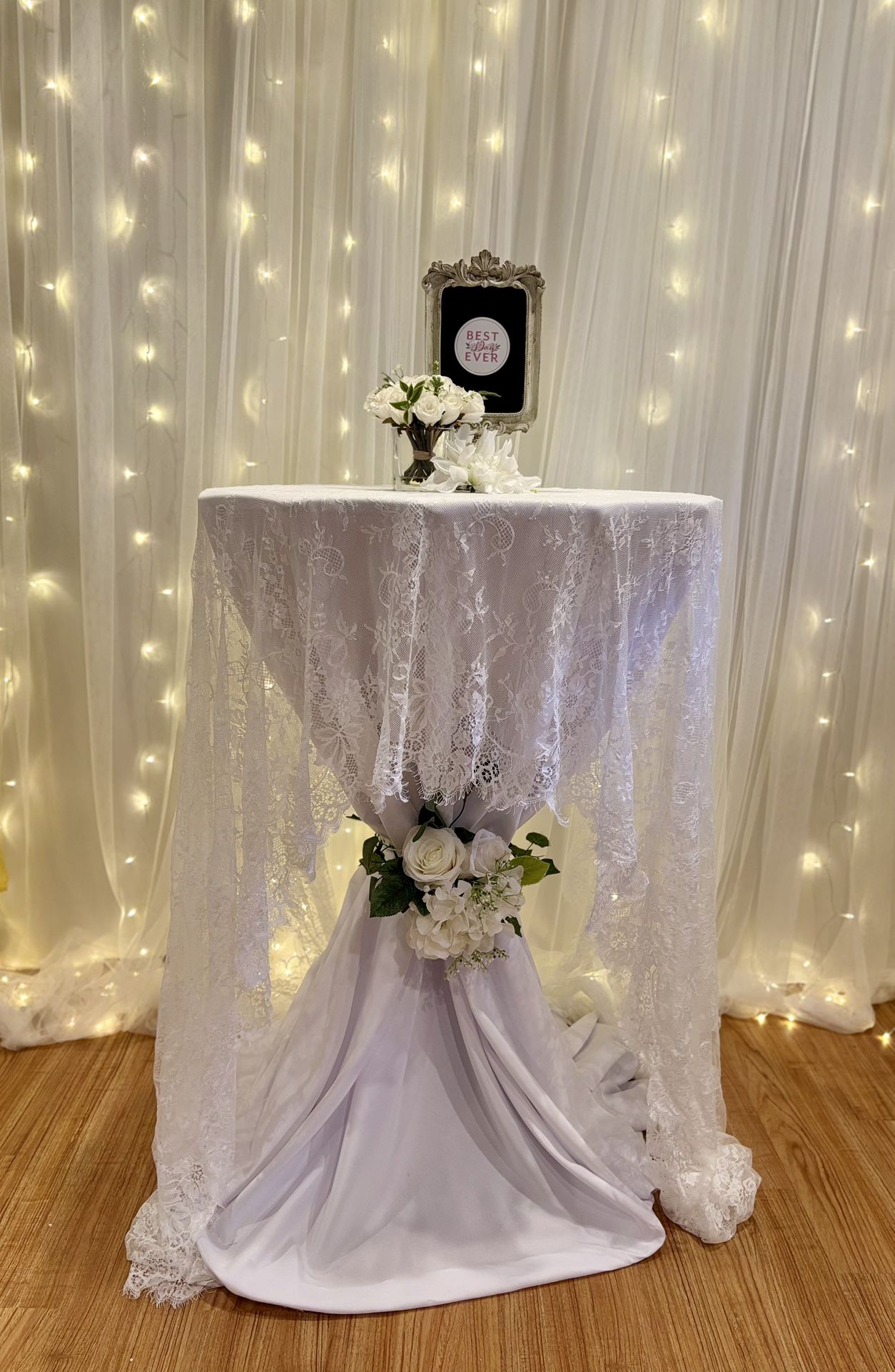 Elegant Wedding/Event Cocktail Table Setup White table cover with delicate lace overlay Stylish floral arrangement with white roses & greenery