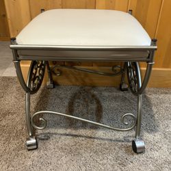 Victorian-style metal vanity stool with a silver finish, off-white upholstered seat, and casters for mobility.  