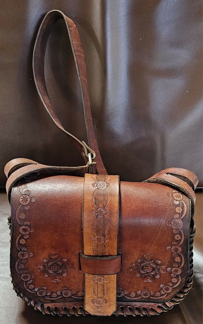 Tooled leather crossbody bag, also known as a saddle bag or satchel. The bag features a floral design that is pressed or carved into the leather.