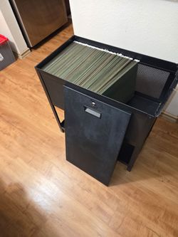 Black metal file cabinet with a flip-top lid and front door, loaded with green hanging file folders. Features a mesh side panel, open bottom shelf, an