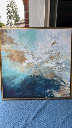 The framed abstract wall art features a dynamic blue, aqua, and gold liquid acrylic composition encased in a slim gold metal frame.