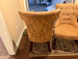 This is a gold upholstered button-tufted wingback dining chair featuring flared sides and dark wooden legs.