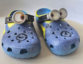 The product is a pair of Crocs Minions clogs in size J3, featuring a slip-on design in a blue color. These casual clogs are made with a synthetic uppe