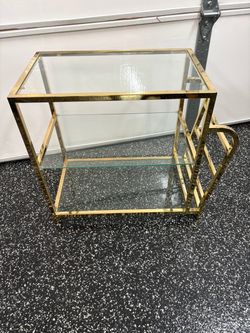 This two-tier brass and glass side table features a sleek, rectangular metallic frame with inset glass shelves and a distinctive curved handle detail