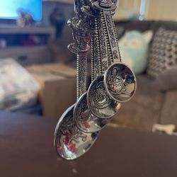 This item is a set of vintage-style ornate pewter measuring spoons featuring a whimsical tea theme. Each spoon is uniquely embossed with detailed desi