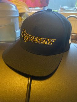 black snapback hat features a gold embroidered "PRESENT" logo on the front and a small rectangular logo on the side.