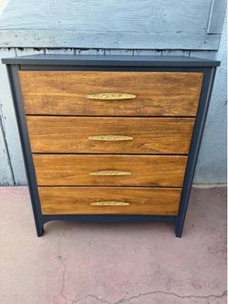 Stylish dresser has a dark gray frame with four wooden drawers. Each drawer has an ornate gold-colored handle. This piece has a mid-century modern des