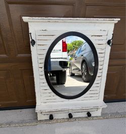 distressed white shutter-style wall mirror features a central oval glass pane in a black frame and built-in hooks with a small display shelf at the bo
