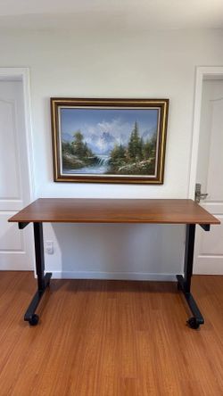 Adjustable standing desk has a wood-grain laminate tabletop and a black metal frame. The height is controlled by a manual hand crank located on the si