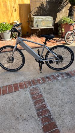 The image shows a GenZe Sport e101 electric bike.  It features a 250-watt geared rear hub motor. The bike has a 7-speed Shimano drivetrain and Promax 