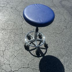  blue rolling stool designed for salon, drafting, or medical use, featuring a vinyl-covered seat, adjustable hydraulic lift, a chrome base, and swivel