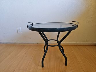 A round, low table with a glass top and a metal frame (also known as an outdoor end table or tray table) with handles.