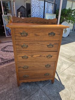 This antique oak chest of drawers features five horizontal drawers, original brass hardware, and a decorative carved crest at the top.