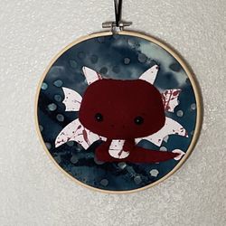 handmade wall hanging featuring a felt Burgundy Axolotl  mounted in an embroidery hoop 