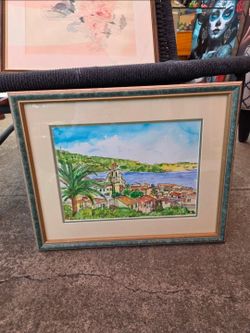 Framed watercolor painting depicting a coastal landscape with red-roofed houses, a palm tree, and a tall tower by the water.