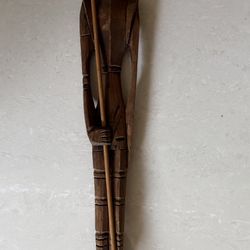 A tall, slender, hand-carved wooden sculpture of a person holding a long staff or spear., $15