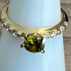 Elegant gold tone ring featuring a vibrant green center stone. Clear accent crystals add sparkle around the band. A refined, eye-catching piece that c