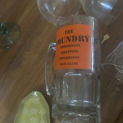 tall glass beer mug from The Foundry Supply Co.  The mug features an orange label with the text "THE FOUNDRY PROVISIONS LIBATIONS DIVERSIONS MUG CLUB"