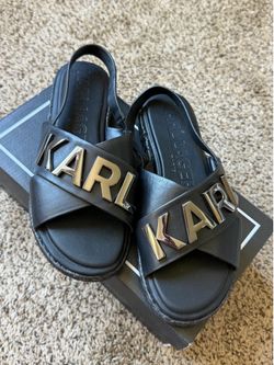 Crisscrossing leather straps across the vamp featuring the large metallic "KARL" logo. Construction: Open-toe design with an elastic or adjustable sli
