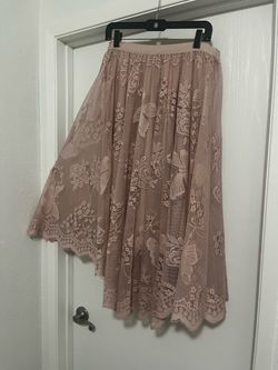 Dusty pink lace midi skirt has an asymmetrical high-low hemline. The fabric features an intricate floral and butterfly pattern with a scalloped edge.