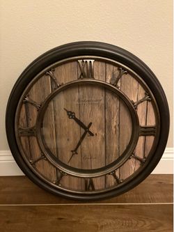 oversized rustic analog wall clock, featuring a distressed faux wood panel face, a dark bronze frame, and raised Roman