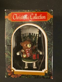 Vintage holiday tree ornament featuring a small brown teddy bear wearing a striped red scarf, sitting in a miniature rocking chair next to a wrapped p