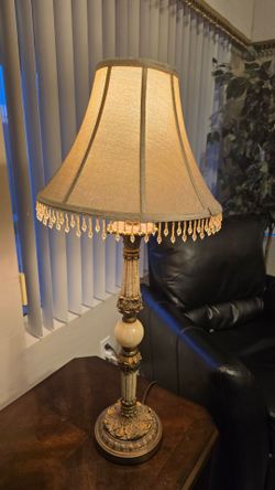 Antique-style table lamp featuring an ornate gold-detailed stand and a fabric shade with hanging beaded fringe.